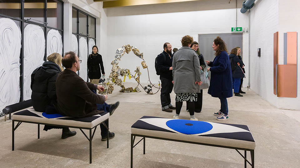 An exhibition with seats with eyes on them and groups of people gathering in the Garcia Family Foundation Gallery at the CCA