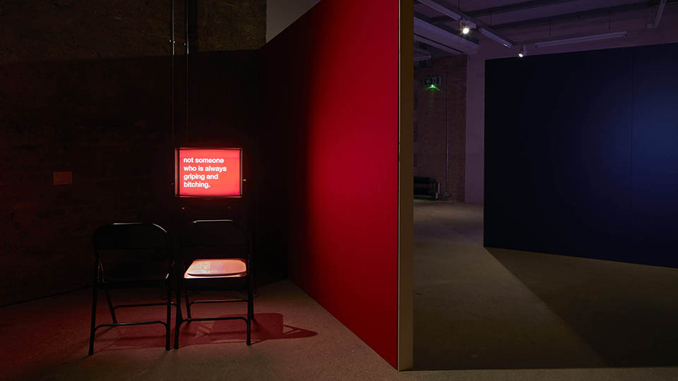 A dark exhibition with a red wall and a screen saying 'not someone who is always griping and bitching' with seats in front of it